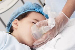 Momentos antes de cirurgias em crianças. Uma pessoa adulta está segurando um aparelho de nebulização no rosto de uma criança.